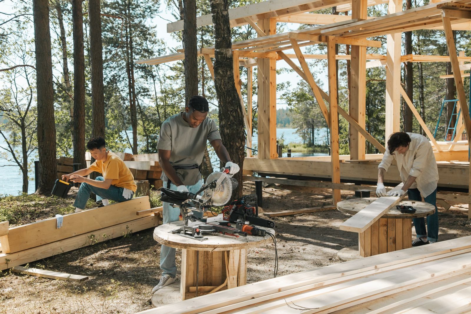 Three carpenters at work on a timber frame in dappled midday sun — one at a mitre saw, one measuring a plank